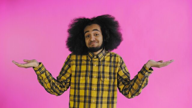 A young man with an African hairstyle on a pink background shrugs his shoulders. Emotions on a colored background.