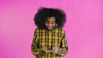 A young man with an African hairstyle on a pink background looks at the phone and happily texts with someone. Emotions on a colored background