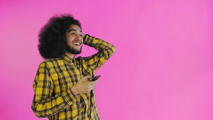 A young man with an African hairstyle on a pink background looks at the phone and is happily surprised. Emotions on a colored background