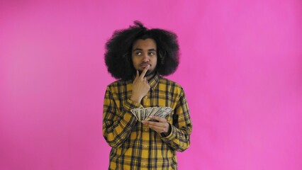 A happy African-American holding money in his hands and thinking about what to spend it on, stands isolated on a pink background