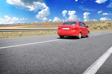 Obraz premium Red car on an asphalt road with fields against a sky wiht clouds