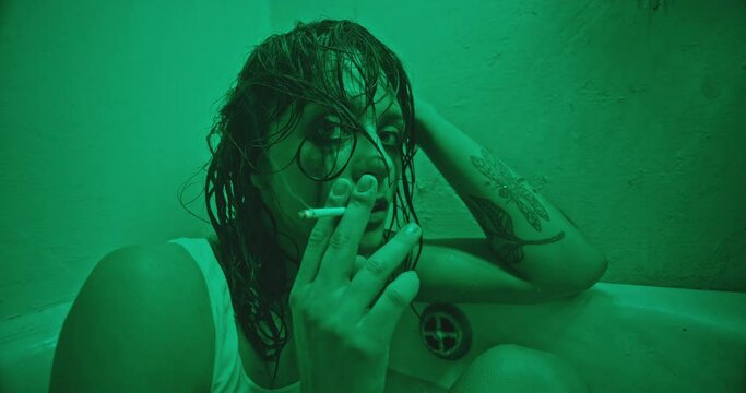 Woman smoking weed in bathtub. Zoom out view of female junkie in wet clothes smoking marijuana joint and looking at camera while sitting in bathtub under green light