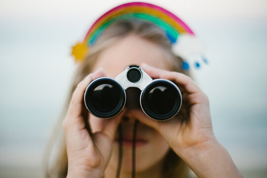 Girl with blond hair and rainbow headband looks through binoculars