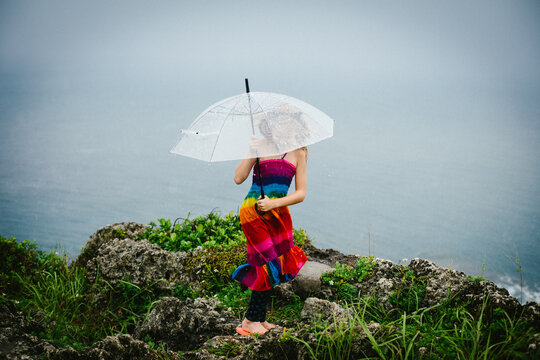 Girl plays in rain with umbrella on cliff by ocean in rainbow dress