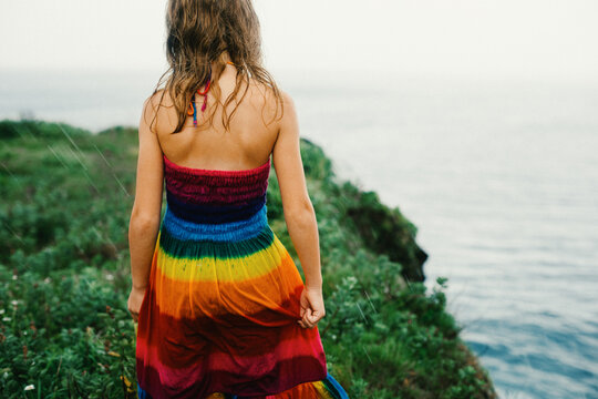 Girl walks in rain on green cliff by ocean in a rainbow dress
