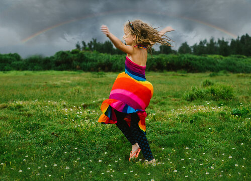 Child blond girl twirls in rainbow dress under a rainbow