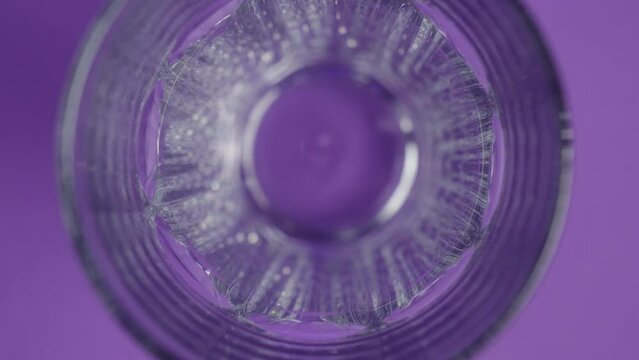 A transparent glass stands on a purple background, the camera is located on top. Ice cubes slowly fill the glass