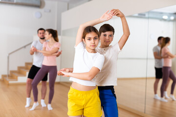 Positive friendly teen sister and brother learning to dance waltz in pair in studio against backdrop of dancing parents. Family bonding and active lifestyle concept..