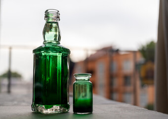 botella grande acompañada de frasco pequeño con liquido con tinta verde diluida con fondo desenfocado de edificio - obrazy, fototapety, plakaty