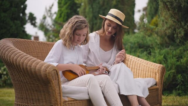 Focused pretty teenage girl learning playing ukulele with young woman teaching sitting on backyard outdoors. Caucasian teenager enjoying weekend with talented sister. Slow motion