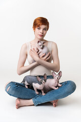 Obraz na płótnie Canvas Redhead young woman holding Sphynx Cat in hands and two kittens sitting on her legs. Hipster woman with short hair in T-shirt and jeans looking at camera, sitting on white background. Part of series