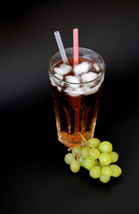 Fresh grape juice in a tall glass with ice and straws on a black background, next to a bunch of ripe white grapes.