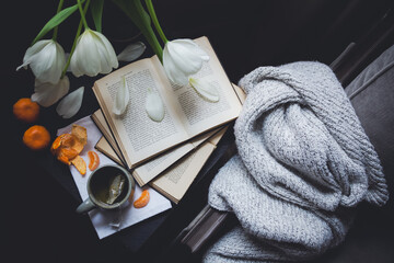 Flat lay of tulips, books, blanket, tangerines, cup of tea on a tabletop next to window on cloudy day