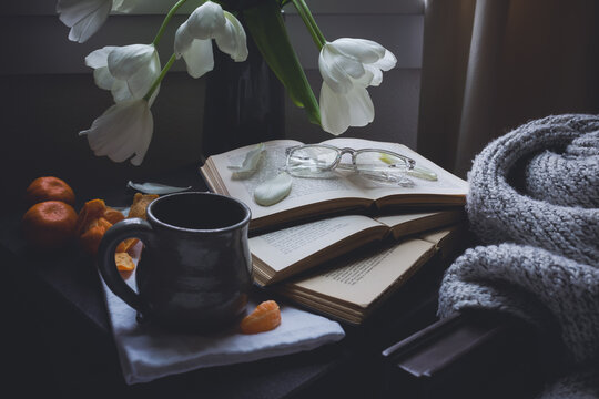 Cup of tea, three antique books with reading glasses, white tulips, peeled tangerines and a blanket next to a window on a cloudy day.