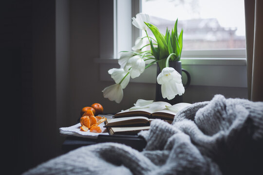 Vase of white tulip flowers with three open books and tangerines on a table next to a window on a rainy day.