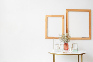 Stylish jewelry, vase with flowers on table and blank frames on light wall