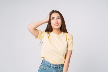 Young unhappy asian woman with long hair in yellow shirt and jeans holding her hands on head on grey background