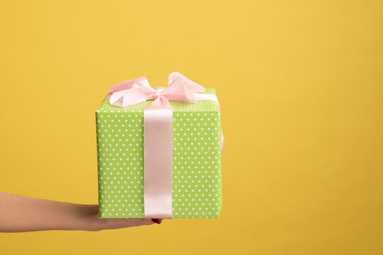 Closeup Side View Of Woman Hand Holding Out Green Gift Box With Ribbon, Giving Present On Holiday, Bonuses And Surprises Concept. Indoor Studio Shot Isolated On Yellow Background.