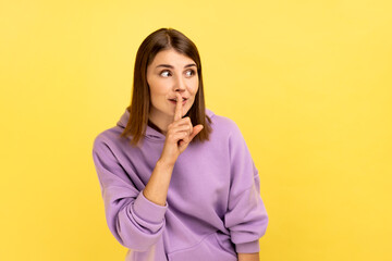 Shh, it's big secret. Beautiful cunning woman smiling, showing gesture secret sign with finger near her lips, looking away, wearing purple hoodie. Indoor studio shot isolated on yellow background.