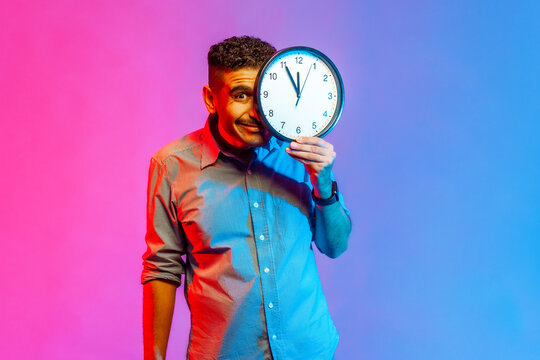 Portrait of funny positive man in shirt standing covering half of face with big wall clock, looking at camera with positive expression. Indoor studio shot isolated on colorful neon light background.