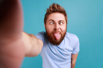Portrait of handsome childish bearded man blogger making point of view phone, showing tongue out,...