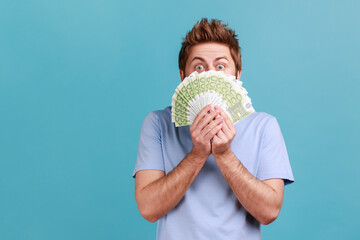 Portrait of handsome young adult bearded man peeping out euro banknotes with playful look, enjoying big jackpot, financial success. Indoor studio shot isolated on blue background. - obrazy, fototapety, plakaty