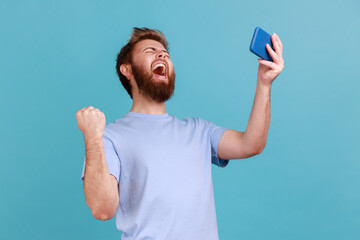 Portrait of extremely happy handsome bearded man gamer playing video game on mobile phone,...