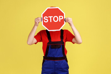 Portrait of worker woman standing hiding her face behind red stop sign, prohibitions and restrictions, wearing overalls and red cap. Indoor studio shot isolated on yellow background.