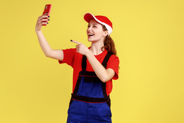 Portrait of happy smiling worker woman broadcasting livestream and pointing finger to mobile phone camera, wearing overalls and red cap. Indoor studio shot isolated on yellow background.