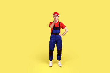 Full length portrait of smiling satisfied woman worker standing and pointing finger to camera, choosing you, wearing overalls and red cap. Indoor studio shot isolated on yellow background.