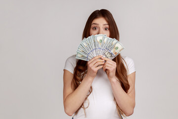 Amazed happy woman with brown hair hiding face behind fan of dollars banknotes, interest-free cash withdrawal, wearing white T-shirt. Indoor studio shot isolated on gray background.
