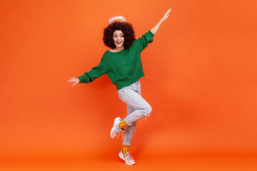 Fototapeta na wymiar Full length of happy excited woman with Afro hairstyle wearing green casual style sweater standing with raised arms, having excited facial expression. Indoor studio shot isolated on orange background.