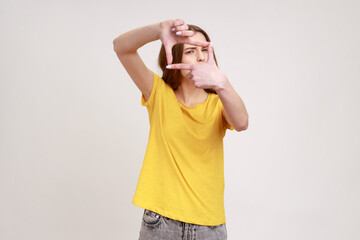 Winsome woman of young age wearing yellow casual T-shirt making frame gesture with her fingers as she looks through camera, composition of photograph. Indoor studio shot isolated on gray background.