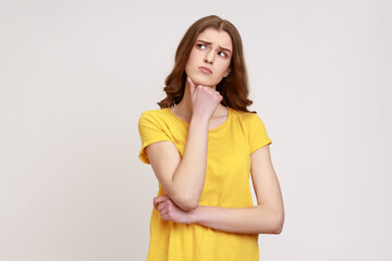 Portrait of attractive teenager girl with brown wavy hair in yellow T-shirt thinking about important things, holding chin, looking away. Indoor studio shot isolated on gray background.