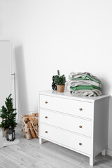 Commode with warm sweaters, small Christmas tree and decor near light wall