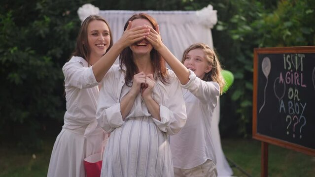 Surprised pregnant Caucasian woman standing on backyard as daughters covering eyes with hands smiling giving gifts for baby shower party. Portrait of excited happy mother enjoying celebration outdoors