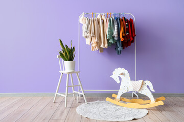 Rack with children sweaters, rocking horse and houseplant near lilac wall