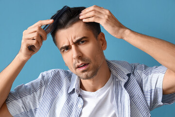 Worried young man combing hair on blue background, closeup