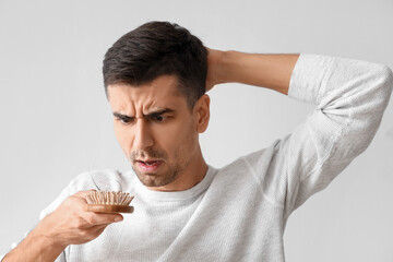 Worried young man holding brush with fallen down hair on light background