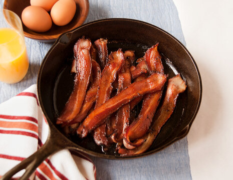 Crispy Bacon Strips in a Cast Iron Skillet; Glass of Orange Juice and a Bowl of Eggs