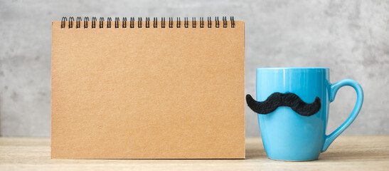 Blue coffee cup or tea mug with Black mustache decor and empty paper notepad or calendar on table....