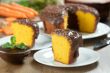 Close up piece of Brazilian carrot cake with chocolate frosting on wooden table with carrots in the background