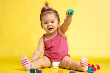 A 1 years old girl is playing with a children's multi-colored constructor. The girl is sitting on a yellow background in a diaper.