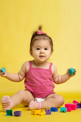 A 1 years old girl is playing with a children's multi-colored constructor. The girl is sitting on a yellow background in a diaper.