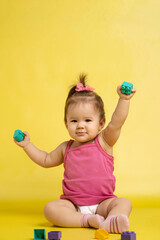 A 1 years old girl is playing with a children's multi-colored constructor. The girl is sitting on a yellow background in a diaper.