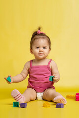 A 1 years old girl is playing with a children's multi-colored constructor. The girl is sitting on a yellow background in a diaper.