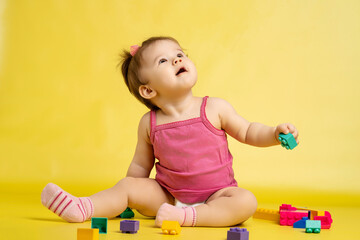 A 1 years old girl is playing with a children's multi-colored constructor. The girl is sitting on a yellow background in a diaper.