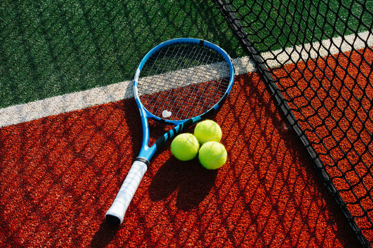 Tennis racket and three standart balls with net shadow cast on it. On a brand new outdoor court. Vibrant green and brown with clear white lines.