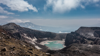 Turquoise acid lake in the crater of an active volcano. Glare on the water. Snowmelt on the banks and on steep slopes.  A mountain on a background of blue sky and clouds. Kamchatka. Gorely Volcano