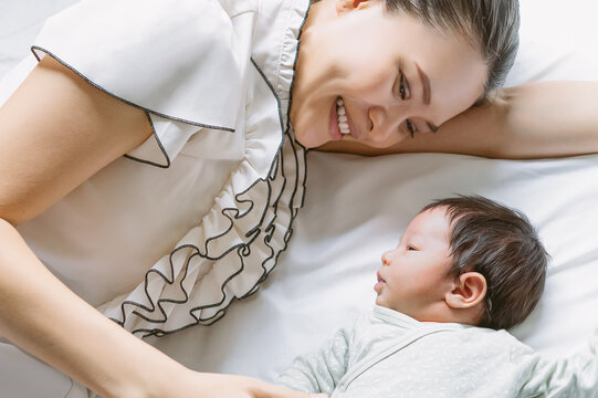 Portrait of young Asian mother and cute newborn baby lying on white bed at home. Maternity concept, soft image of beautiful family, mother's day concept.